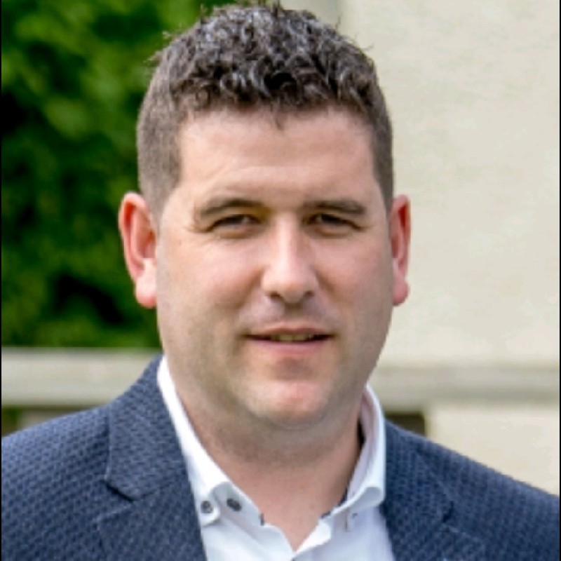 John Roulstone - Fresh Thinking Labs Portrait of a man in a dark blue blazer and white shirt, smiling slightly, outdoors with greenery in the background