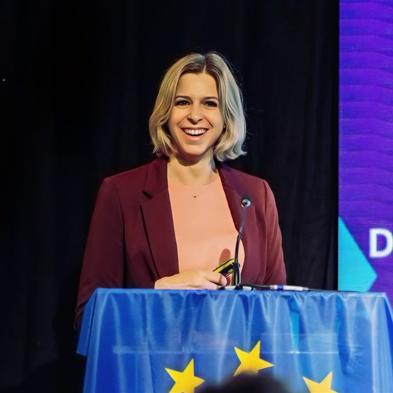 Martina Wolfgruber - Fresh Thinking Labs Smiling woman in a burgundy blazer and peach top speaks at a podium with a blue cloth and yellow stars, microphone in front of her.