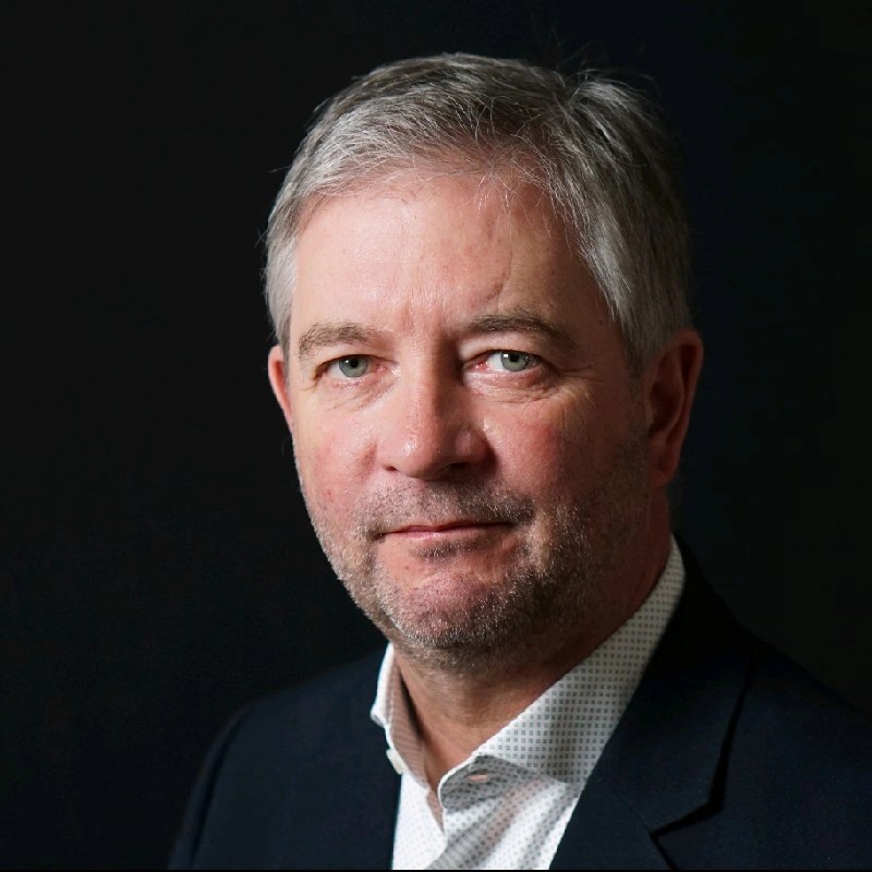 Professional head-and-shoulders portrait of a middle-aged man with gray hair in a dark blazer against a dark background.
