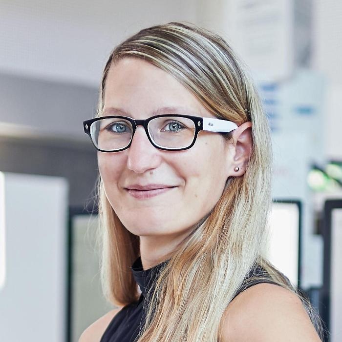 Woman with blonde hair and black-rimmed glasses smiling in an office setting with computers blurred in the background
