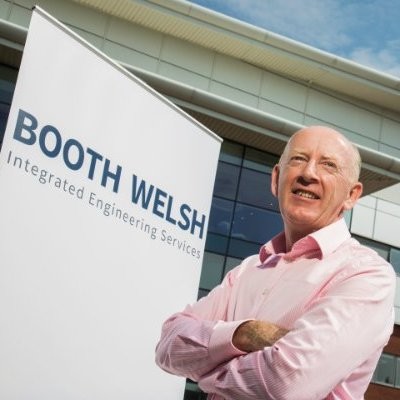 Martin Welsh - Fresh Thinking Labs Smiling man in a pink shirt stands beside a Booth Welsh Integrated Engineering Services sign outside a modern building.
