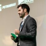 Simon_Winter - Fresh Thinking Labs Man in a dark suit giving a talk on stage, holding a green card and wearing a headset microphone.