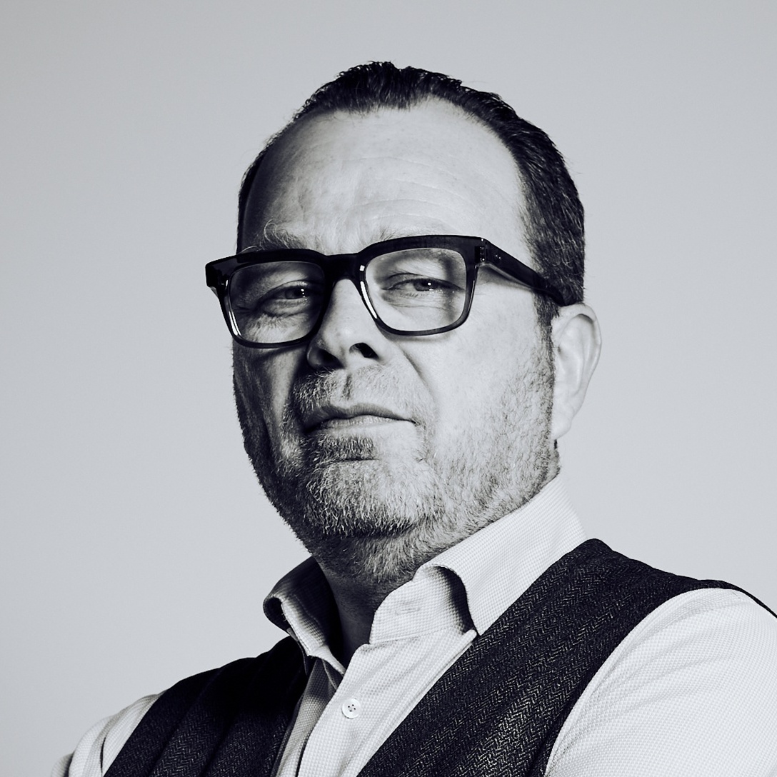 Portrait of a middle-aged man wearing dark-framed glasses, a light button-down shirt, and a dark vest, looking slightly to the left.