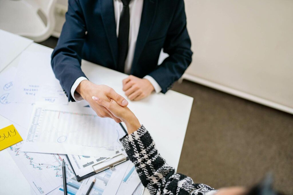 Two professionals shake hands over a desk crowded with charts and documents, signaling a business agreement.